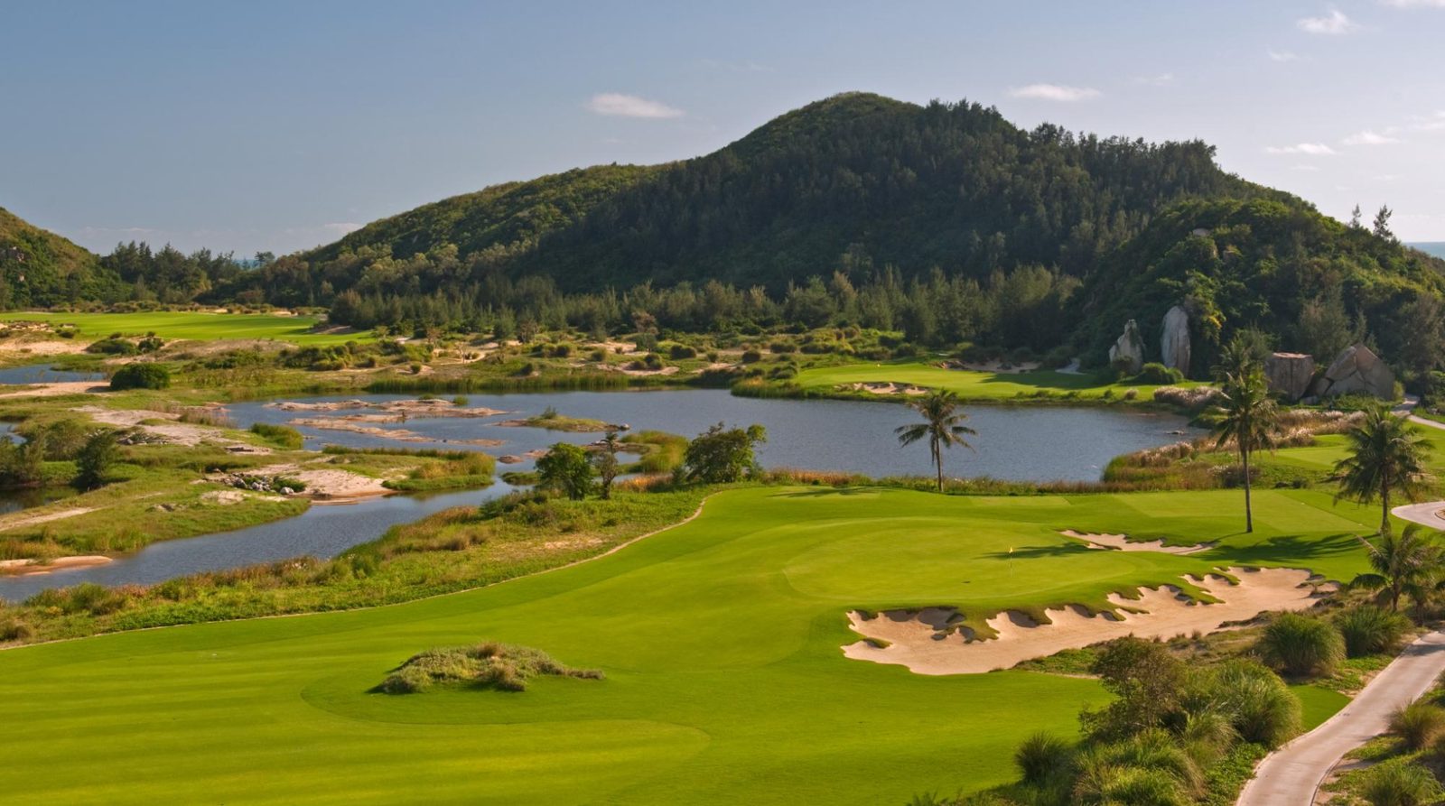 A green beside a lagoon with a forested hill behind at The Dunes, Shenzhou Peninsula