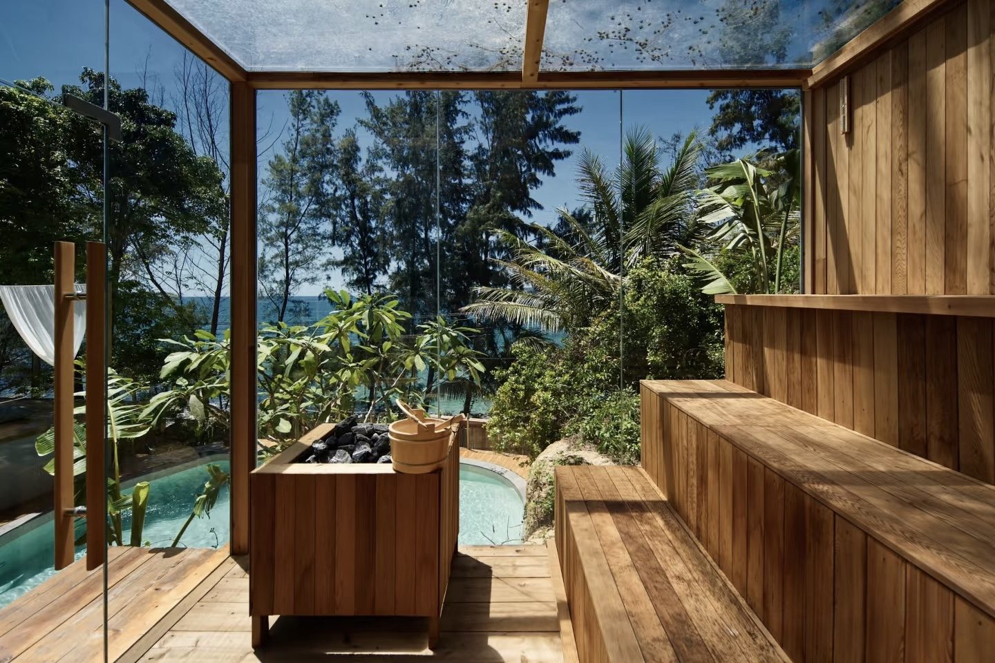 Glass-walled wooden sauna interior looking out to tropical trees and the sea at the recovery space near Riyue Bay, Wanning, Hainan — sauna after surfing